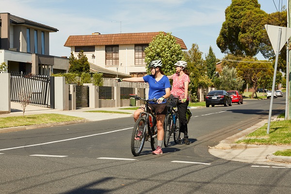 two adult bike riders waiting at an intersection, both signalling their intention to turn right by holding their right arms out to their side.