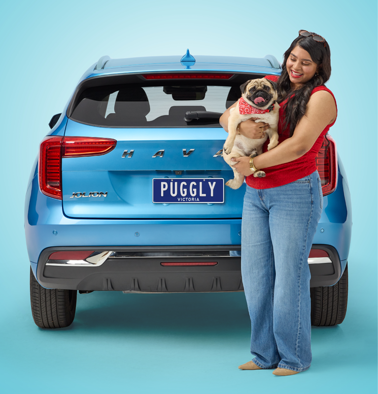 Woman with pug dog stands in front of blue car featuring the number plate 'Puggly'.