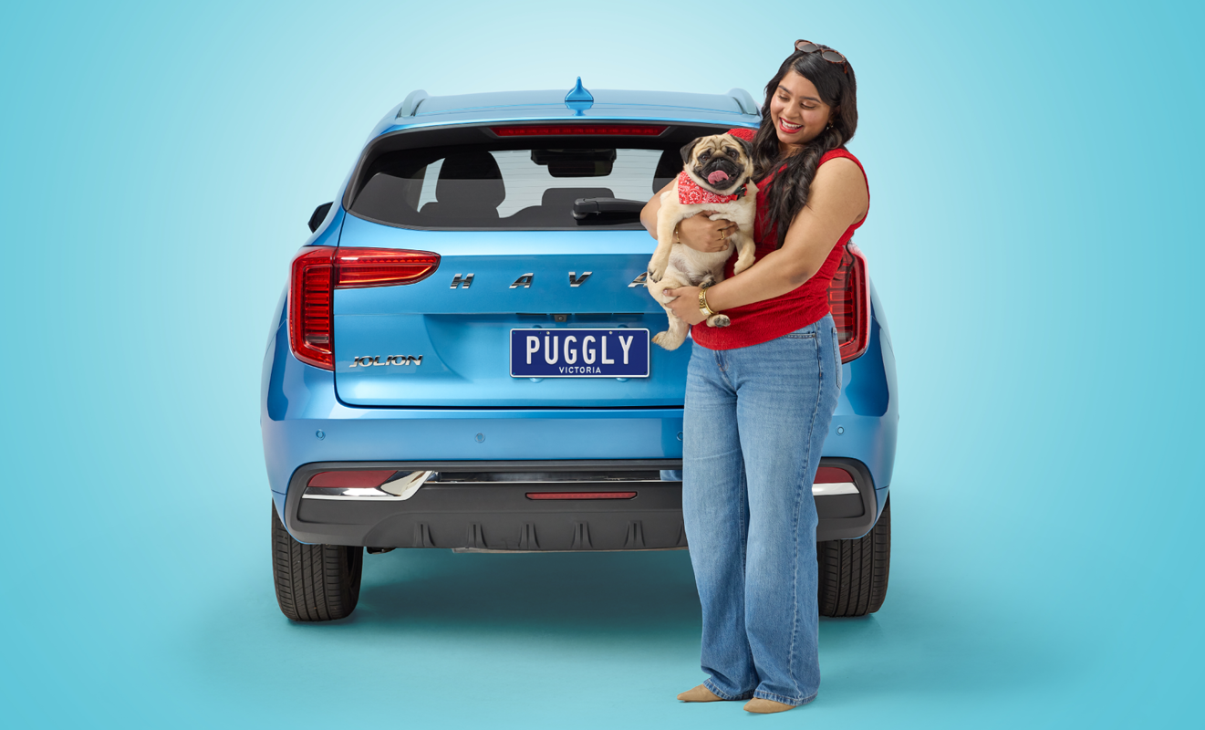 Woman with pug dog stands in front of blue car featuring the number plate 'Puggly'.